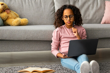 Remote education. Thoughtful black gen z girl sitting on floor, using laptop and looking at book. Schoolgirl wearing eyeglasses watching online course, taking notes, doing homework
