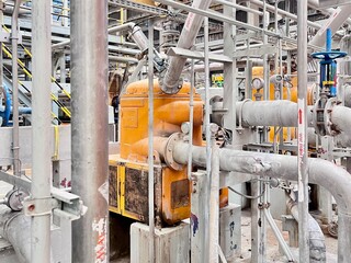 An intricate network of industrial pipes and machinery in a factory setting. A large yellow tank is...