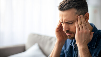 Closeup of middle-aged bearded man suffering from headache at home, touching his temples, panorama...