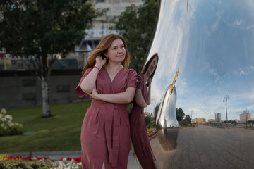 Woman in a rose dress stands by a shiny mirrored sculpture in a city park, her reflection blending...
