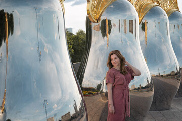 Woman in a rose dress poses beside giant reflective matryoshka sculptures with golden tops on Taras...