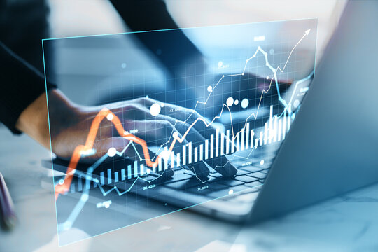 Businessperson analyzing financial market data with virtual hologram charts and graphs displayed over laptop keyboard in digital workspace.