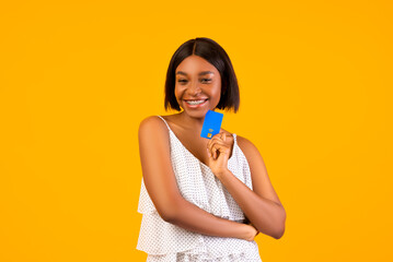 Elegant black woman in summer dress holding credit card, smiling at camera on orange studio background. Charming African American lady planning budget for tropical beach vacation