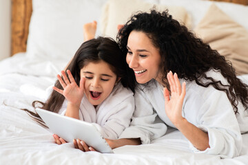 Relaxed mother and little daghter resting at spa, wearing white bathrobes and laying on huge bed, calling their father from hotel room, cheefully waving at digital tablet screen, copy space