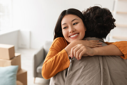 Smiling young Asian woman hugging her boyfriend and holding house key on moving day, free space. Loving millennial couple embracing in their new home during relocation