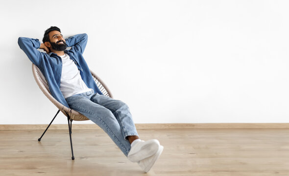 Relaxed young Indian man relaxing with hands behind head in armchair indoor, enjoying comfort and rest on chair near white studio wall. Panorama with empty space for text advertisement