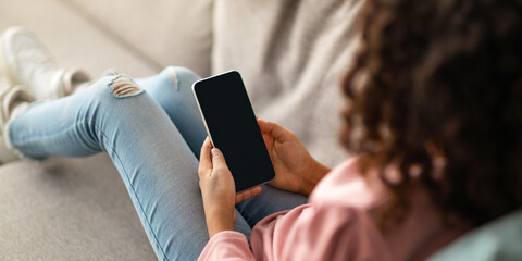 Unrecognizable curly black girl holding smartphone with black blank screen, using mobile application or useful website on sofa. Mockup, cropped, selective focus