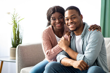 Fototapeta premium Beautiful black couple spending time together at home while COVID-19 pandemic, happy african american young family sitting on couch in living room, embracing and looking at camera, copy space