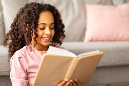 Happy african american teen girl reading paper book, resting at home while sitting on floor near sofa. Little reader spending time on weekend, copy space