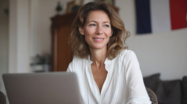 Middle-aged French teacher sits at desk with laptop, ready for an online lesson, with French flag in background, representing remote education and language learning.