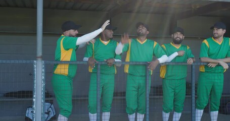 Diverse male teammates clapping in dugout behind metal railing, celebrating teammate raising arms
