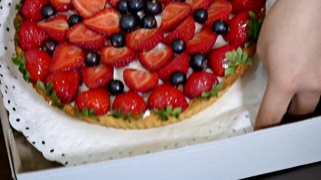 This close-up video showcases a beautifully decorated berry tart, brimming with vibrant strawberries and dark berries. The dessert is artfully arranged on a white doily within a cardboard box
