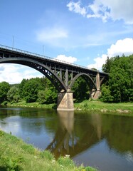Fototapeta premium A dark metal bridge spans a calm river, reflected in its still waters, set against a backdrop of lush green trees and a partly cloudy blue sky