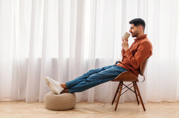 Rest Concept. Happy Arab guy drinking coffee sitting on chair in living room. Casual man relaxing,...