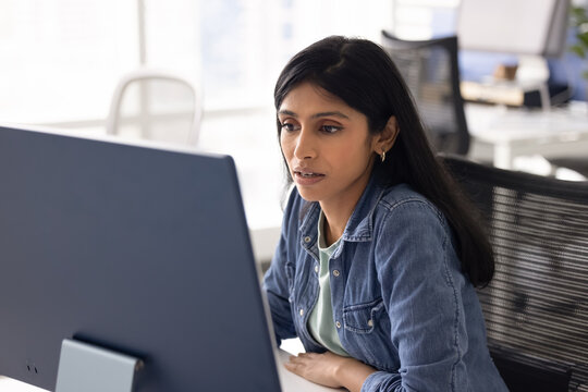 Serious young Indian project manager woman working at computer monitor, using business application on pc for online professional communication, reading or watching content