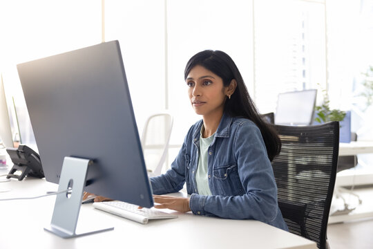 Focused young Indian specialist woman working at computer monitor in office open space alone, using Internet technology, professional application for job task, typing on keyboard