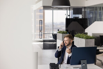 Happy successful business company employee man working in contemporary office workspace, sitting at workplace table, talking on wired telephone, smiling, laughing. Wide candid shot with copy space © fizkes
