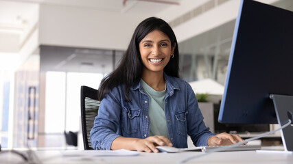 Happy young Indian professional woman posing at office workplace, sitting at computer monitor, working on Internet startup project, looking at camera, smiling, laughing. Banner shot