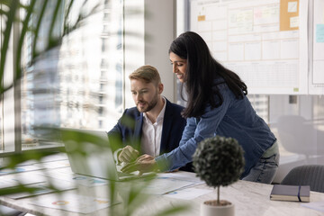 Diverse couple of business colleagues working on Internet project at laptop together, analyzing online sales statistic result. Manager giving professional help, support to coworker. Candid shot