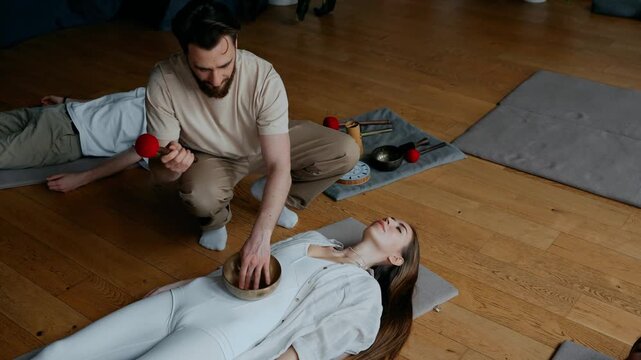 A male yoga mentor hits a Tibetan bowl with a mallet on the stomach of a female student who is lying on the mat with her eyes closed