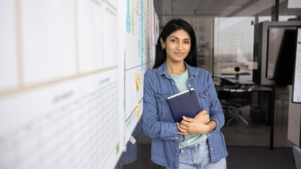 Positive young Indian company director woman in casual denim clothes posing at white board in office workspace, standing with hands folded, looking at camera with friendly smile. Banner shot