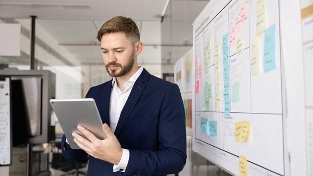 Serious young company CEO man in formal suit browsing business social media on digital tablet in office, using gadget, modern electronic technology for job communication, project plan. Banner