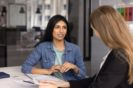 Two young diverse company colleagues talking at workplace office table, discussing project marketing strategy. Indian manager woman asking expert, consultant for advice