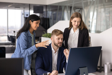 Diverse team of three coworkers discussing online product at monitor, speaking at workplace,...
