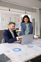 Two positive diverse millennial colleagues analyzing online marketing sales, Internet statistic reports at laptop, watching funny content, pointing at screen, smiling, laughing. Vertical shot