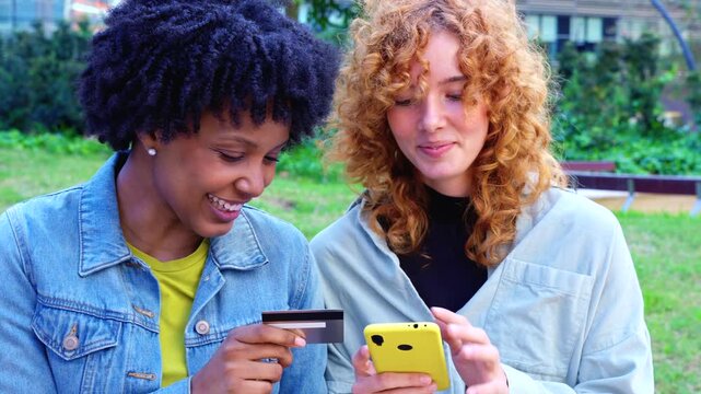 Cheerful African American and Caucasian friends stand in a city park, holding colorful smartphones and a credit card, celebrating cashless convenience, shared shopping success, and digital lifestyle