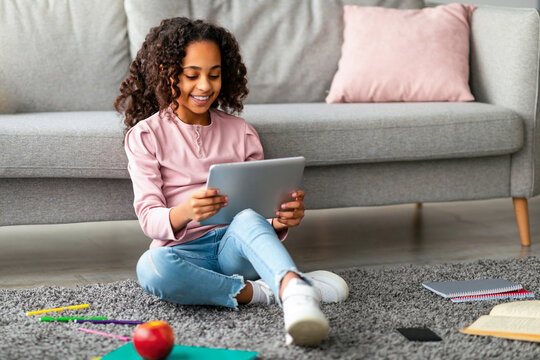 Smiling teen african american girl using modern digital tablet at home, sitting on floor carpet with books and watching online lesson on pad, using application for e-education, copy space