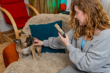 Young Caucasian redhead woman smiles while using smartphone to take photo of pug dog sitting on...