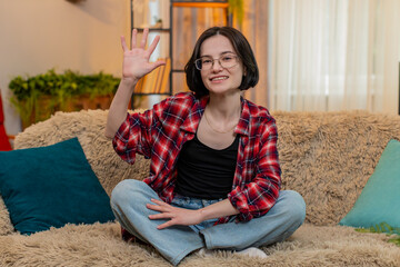 Young woman smiles waves hand hello at home while sitting on sofa happily greeting someone with joy...
