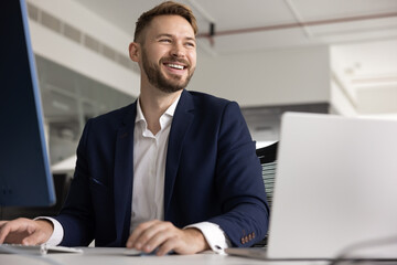 Positive handsome young Caucasian professional man working at laptop, using large monitor, looking away, smiling, laughing. Happy successful legal expert, CEO, executive candid portrait