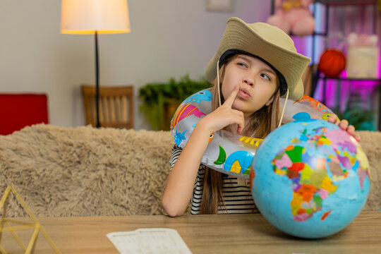 Young child girl sitting at home table wears a travel hat and inflatable ring while looking at a globe, imagining a beach vacation. School kid enjoys dreaming about travel planning a seaside holiday