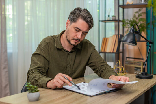 Middle-aged man at home reads contract document thinking deeply weighing decision feeling cautious. Young adult guy at table taps pen hesitates over legal terms before signing important agreement now - Powered by Adobe