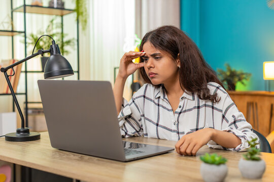 Indian woman at home holds head staring at laptop baffled by complex task feeling stressed missing answer. Arabian girl at table shows confusion scrolling files on desk trying find solution quickly.