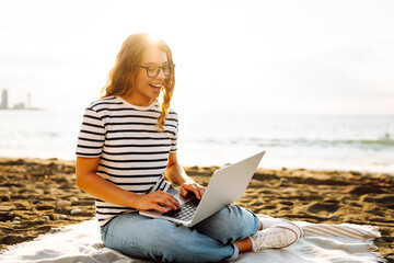 Young woman sitting on mat with laptop on sunset beach. Female freelancer in casual clothes...
