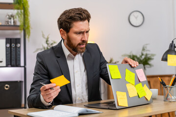 Middle-aged businessman at home office looks at laptop full of stickers for tasks and feels pressure. Freelancer guy at table worries about deadlines types fast and tries to finish plan before end