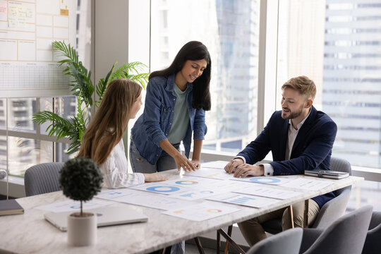 Positive diverse team of three project managers discussing marketing strategy, finance, investment for startup at large office table, analyzing paper sales reports, sharing ideas, insights