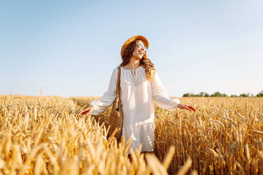 Beautiful woman in a hat walks in a golden wheat field at sunset. Happy woman in a white dress enjoys a rural landscape. Beauty, fashion, weekend concept. Lifestyle.