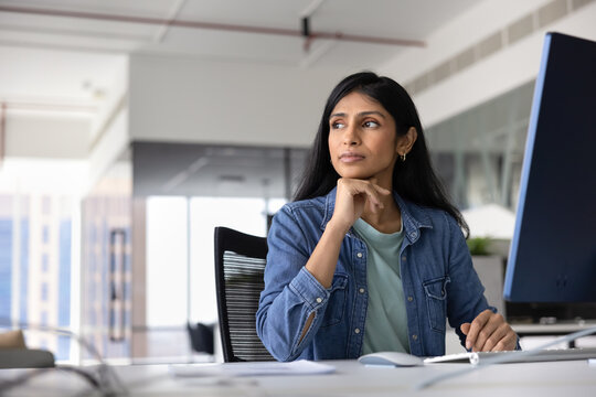 Thoughtful successful young female Indian company worker in casual sitting at pc monitor at workplace, looking away, thinking on job task, creative idea for startup, planning strategy
