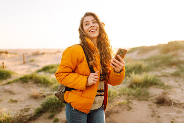 Portrait of female hiker with phone on path leading to beach. Young woman enjoying sunrise and texting on smartphone with seascape. Adventure and technology concept.