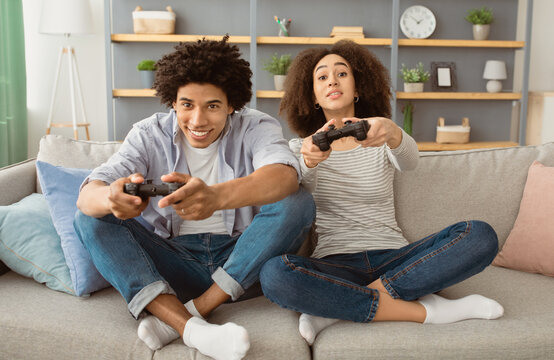 Emotions from online games during self-isolation of covid-19 and entertainment at home. Excited young african american male and female playing with joysticks sitting on sofa in living room interior