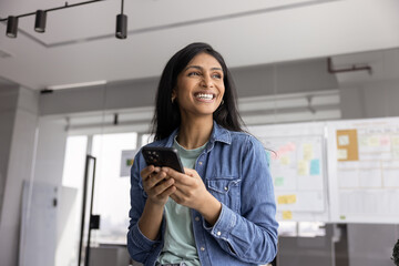 Cheerful young Indian company employee in casual clothes typing on smartphone in office on work...