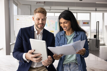 Cheerful young multiethnic coworkers comparing data on tablet and in paper reports, watching online content on digital gadget, holding stack of documents, talking in office, smiling, laughing