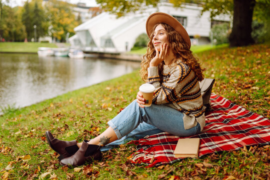 Cute woman in hat relaxing on river bank sitting on red blanket with coffee cup. Beautiful woman enjoying autumn weather in park with drink. Relaxation, weekend concept. - Powered by Adobe