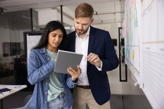 Diverse couple of young business colleagues using online application for project management tablet computer, talking at scrum board with sticky notes, discussing project plan, tasks scheduling