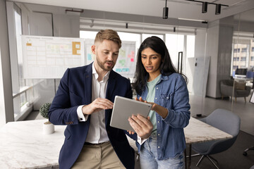 Young Caucasian business man showing tablet to female Indian coworker, sharing ideas for online...