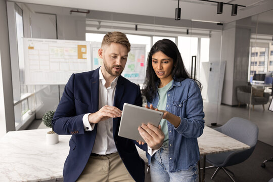 Young Caucasian business man showing tablet to female Indian coworker, sharing ideas for online project, asking for advice, professional help, support. Diverse colleagues using gadget for teamwork
